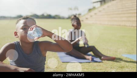 Bild von Lichtflecken über glücklichen, vielfältigen Schülern, die Wasser in Outdoor-Yoga-Klassen trinken Stockfoto