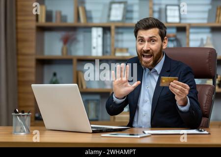 Ein aufgeregter reifer Geschäftsmann in einem Anzug macht mit einer Kreditkarte am Schreibtisch im Home Office eine unerwartete Entdeckung. Stockfoto