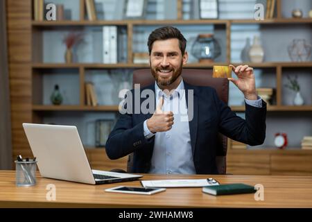 Lächelnder reifer Geschäftsmann in einem eleganten Anzug, der eine Kreditkarte vorlegt, während er von einem ordentlichen Heimbüro aus arbeitet. Stockfoto