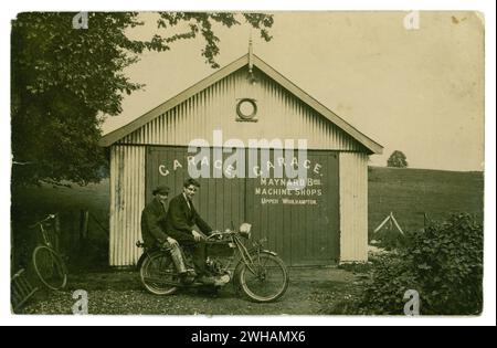 Ursprüngliche Postkarte aus den frühen 1900er Jahren von zwei jungen Jungs, Freunden, die zusammen auf einem Motorrad vor der Maynard Brothers Garage, Machine Shops, Upper Woolhampton, Berkshire, Großbritannien sitzen. Ein Fahrrad steht draußen. Einer der Jungs trägt improvisierte Schutzbrillen, der Sozius trägt lackierte Hosen von Arbeitern. Um die 1920er Jahre Stockfoto