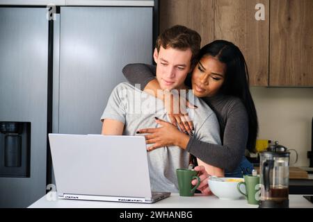 Multiethnisches Paar, Mann arbeitet, während seine Frau ihn umarmt. Stockfoto