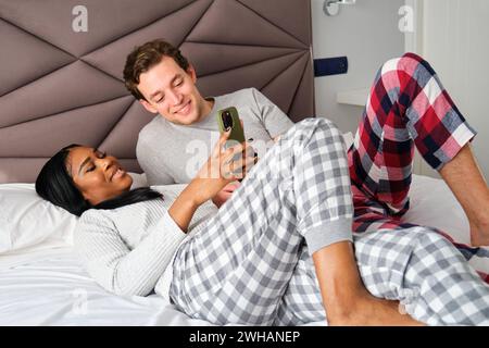Multirassisches Paar benutzt das Telefon zusammen im Bett. Stockfoto