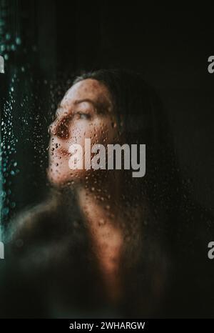 Frauengesicht hinter einer Fensterscheibe, bedeckt mit Regentropfen. Stockfoto