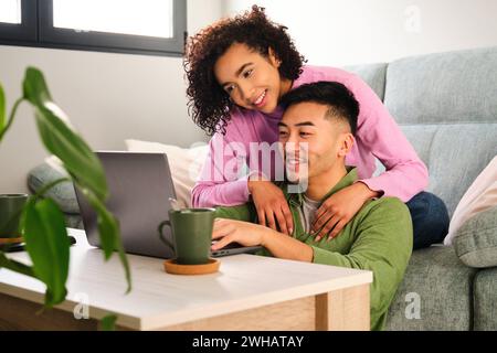 Multirassisches glückliches Paar, das zu Hause den Laptop auf einem Sofa benutzt. Stockfoto