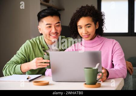 Multiethnisches Paar lächelt, benutzt den Laptop und trinkt Kaffee. Stockfoto