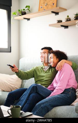 Multirassische Paare lächeln und sehen gemeinsam Fernsehen, Filme oder Filme auf dem Sofa. Stockfoto