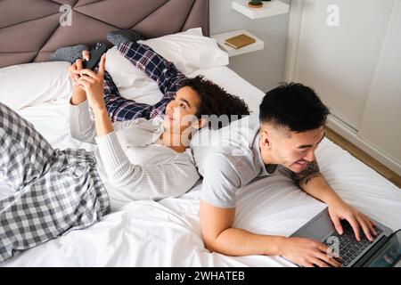 Vielseitiges glückliches Paar, das Smartphone und Laptop zusammen im Bett nutzt. Stockfoto