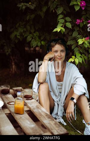 Frau in Kopfhörern trinkt Kaffee, Café, sitzt auf dem Rasen. Bali. Stockfoto