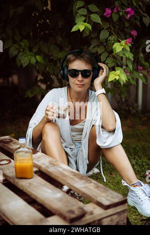 Frau in Kopfhörern trinkt Kaffee, Café, sitzt auf dem Rasen. Bali. Stockfoto