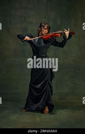 Dynamische Pose einer leidenschaftlichen Musikerin, jungen Frau, Geigerin in schwarzer Kleidung, die mit intensivem Ausdruck auf Vintage-grünem Hintergrund Geige spielt Stockfoto