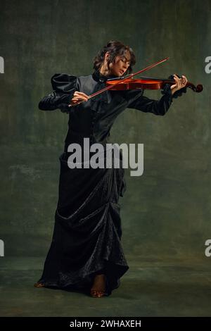 Dynamische Pose einer leidenschaftlichen Musikerin, jungen Frau, Geigerin in schwarzer Kleidung, die mit intensivem Ausdruck auf Vintage-grünem Hintergrund Geige spielt Stockfoto