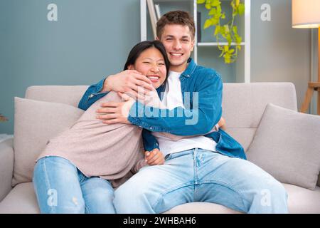 Glückliches junges multirassisches Paar, das sich auf dem Sofa im Wohnzimmer umarmt. Stockfoto