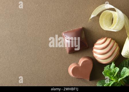 Makroansicht von drei verschiedenen Schokoladenbonbons auf braunem, strukturiertem Tisch mit Goldband und Minzblättern. Draufsicht. Stockfoto