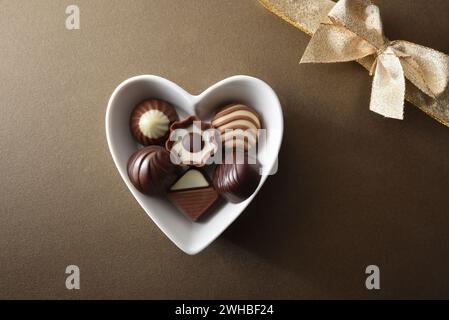 Verschiedene Schokoladenbonbons in weißem Keramikbehälter in Herzform auf braunem strukturiertem Hintergrund mit goldener Geschenkschleife. Draufsicht. Stockfoto