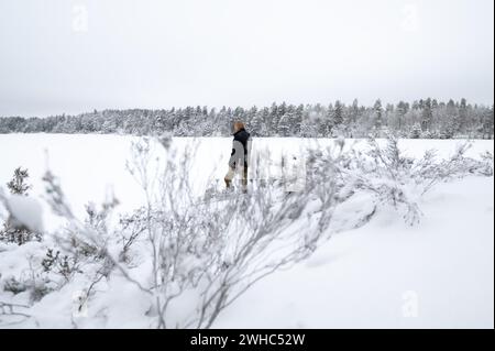 Erwachsener Mann, der am schneebedeckten Seeufer steht und weit weg auf Waldbäume blickt. Verbringen Sie Zeit allein an einem weißen, kalten, schneebedeckten Wintertag. Friedliches atmos Stockfoto