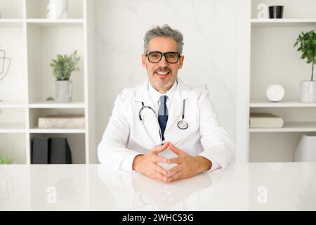 Ein freundlicher Oberarzt, der direkt in die Kamera blickt, die Hände in einer einladenden Geste offen hält und Vertrauen und Kommunikation in einem modernen Gesundheitsumfeld einlädt. Porträt eines erfahrenen Arztes am Arbeitsplatz Stockfoto