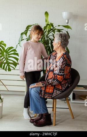 In einem sanft beleuchteten Raum steht ein junges Mädchen neben einer Topfpflanze und teilt einen Moment der Verbindung und Unterhaltung mit ihrer älteren Großmutter, die Freude Stockfoto