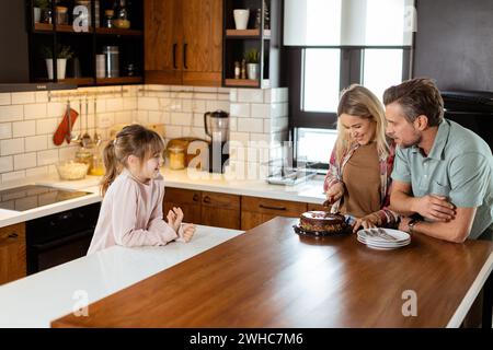 Eine herzerwärmende Szene entfaltet sich, während eine Familie einen köstlichen Schokoladenkuchen zusammen in der Wärme ihrer sonnendurchfluteten Küche genießt und Lächeln und cr teilt Stockfoto