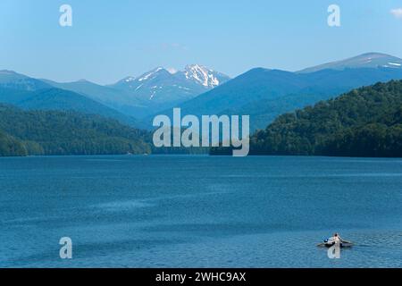 Ein kleines Boot auf einem ruhigen blauen Reservoir mit schneebedeckten Bergen im Hintergrund, Vidraru Reservoir, Transfogarasan High Road, Transfagarasan Stockfoto