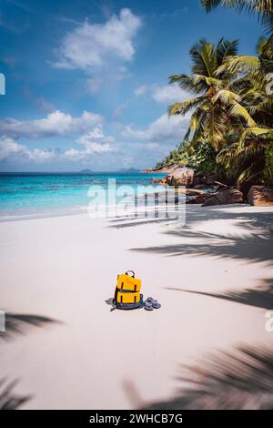 Gelber Rucksack und Flip Flops am wunderschönen tropischen Sandstrand. Palmen und blauer Himmel. Urlaubskonzept. Stockfoto