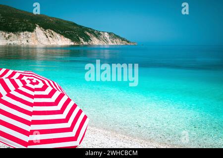 Gestreifter roter Sonnenschirm an einem Kiesstrand mit türkisblauem Meer, weißen Felsen und Himmel im Hintergrund. Stockfoto