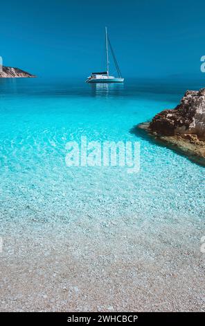 Einsames weißes Segelkatamaran driftet auf ruhiger Meeresoberfläche. Reine azurblaue Lagune mit flachem Wasser und Kieselstrand. Ein paar braune Steinsteine im Vordergrund. Stockfoto