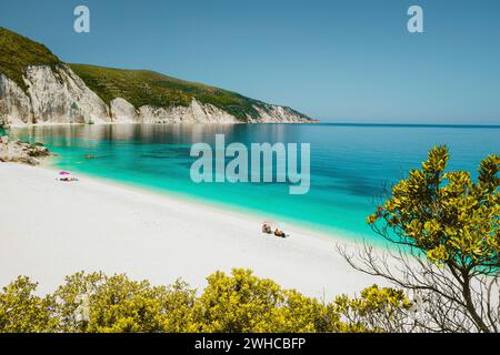 Fantastische Lagune am Strand von Fteri, Cephalonia Kefalonia, Griechenland. Touristen entspannen sich unter dem Sonnenschirm in der Nähe des klaren blauen smaragdtürkisfarbenen Meerwassers. Weiße Steine im Hintergrund. Stockfoto