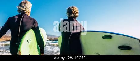 Ein paar Senioren am Strand mit schwarzen Neoprenanzügen Ein Surftisch bereit zum Surfen am Strand - Aktive reife und pensionierte Menschen tun glückliche Aktivität zusammen in Ihre Ferien oder Freizeit Stockfoto