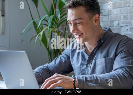 Schöner junger Geschäftsmann mit Brille, der mit dem Computer aus der Ferne arbeitet, am Holztisch im Büro sitzt. Angenehmer glücklicher Mann, der in sozialen Netzwerken kommuniziert, online nach Informationen sucht. Stockfoto