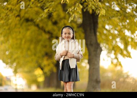 Kindertag. Fröhliches kleines Mädchen 5-6 Jahre alt posiert und lächelt draußen vor der Kamera und steht am Baum im Herbstpark. Stilvoller Zartpressdruck Stockfoto