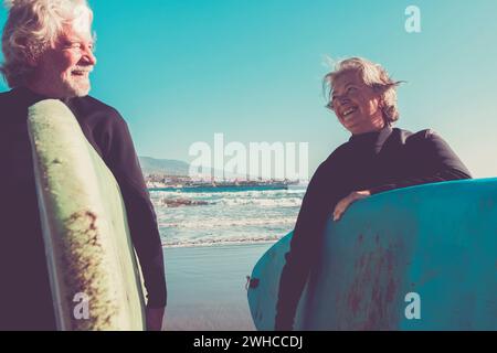 Ein paar Senioren am Strand mit schwarzen Neoprenanzügen Ein Surftisch bereit zum Surfen am Strand - Aktive reife und pensionierte Menschen tun glückliche Aktivität zusammen in Ihre Ferien oder Freizeit Stockfoto