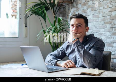 Nachdenklicher Geschäftsmann, der das Kinn berührt, Ideen oder Strategien nachdenkt, mit Laptop am Holzarbeitstisch sitzt, freiberuflich an einem Online-Projekt arbeitet, Student, der sich auf die Prüfung zuhause vorbereitet Stockfoto