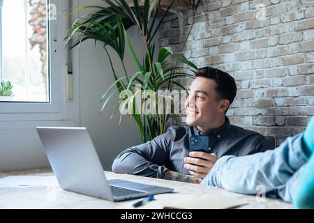 Zufriedener Teenager, der bei der Arbeit mit einem Laptop im Büro gerne telefoniert, die Hände hebt und die Füße auf den Tisch legt, sich nach einem harten Arbeitstag entspannt, in Erwartung eines Wochenendurlaubs, eines entspannten Arbeitstages, ohne Stress Stockfoto