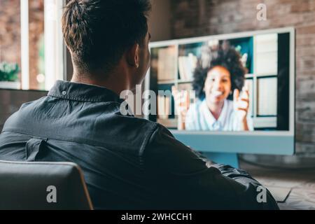 Blick über die Schulter Arbeitgeber Zuhören Bewerber beim Bewerbungsgespräch online mit Kamera und pc. Junger kaukasischer Mann mit Tätowierungen zu Hause, der online mit einer afrikanischen Kollegin im Video arbeitet und spricht. Stockfoto