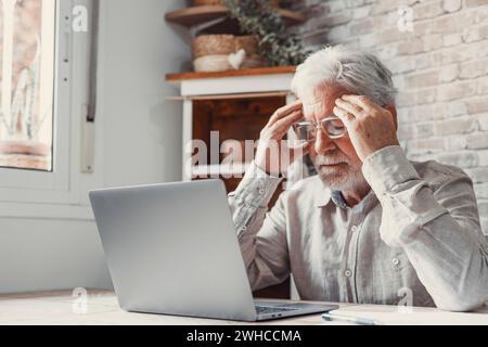 Müder reifer Mann aus den 70er Jahren, der von zu Hause aus am Laptop arbeitet, Brillen abnimmt, Kopf und Augen berührt. Alter Rentner, der an Kopfschmerzen, Schwindel leidet, nachdem er auf den Computer geschaut hat. Sehstörungen Stockfoto