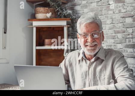 Veralteter Remote-Mitarbeiter. Konzentrierter älterer Mann mit Brille arbeitet am Laptop vom Home Office aus und schaut in die Kamera. Alter Mann Angestellter Freiberufler sitzen am Schreibtisch durch pc-Tippbericht online Stockfoto