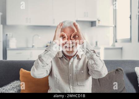 Kopffoto und Porträt eines alten, reifen Mannes, der auf dem Sofa sitzt und in die Kamera schaut und lustige Gesichter mit lächelnden und lachenden Fingern und Händen macht. Gesunde Senioren genießen zu Hause Stockfoto