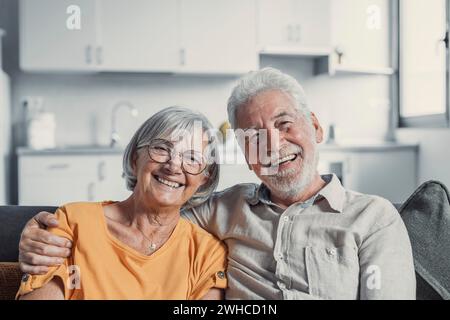 Kopffoto Porträt lächelnder älterer 60er Jahre Mann und Frau sitzen auf der Couch, umarmen Kuscheln, glückliches reifes altes Paar ruhen auf Sofa im Wohnzimmer umarmen Blick auf Kamera zeigen Liebe und Sorgfalt Stockfoto