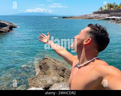 Ein attraktiver und glücklicher junger Mann am Strand, der lächelt und auf das Meer blickt. Die Kamera halten und ein Selfie mit seinem Handy machen. Sommerzeit im Urlaub Spaß und Genießen Stockfoto