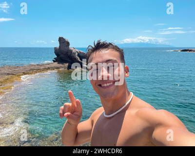 Ein attraktiver und glücklicher junger Mann am Strand, der lächelt und in die Kamera blickt und ein Selfie mit seinem Handy macht. Sommerzeit im Urlaub Spaß und Genießen Stockfoto