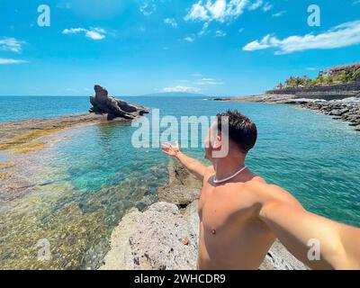Ein attraktiver und glücklicher junger Mann am Strand, der lächelt und auf das Meer blickt. Die Kamera halten und ein Selfie mit seinem Handy machen. Sommerzeit im Urlaub Spaß und Genießen Stockfoto