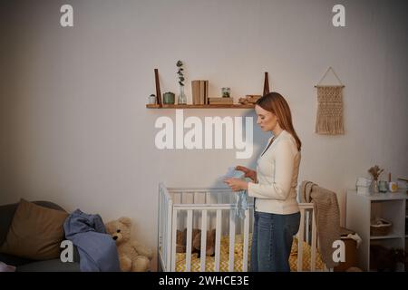 Seitenansicht einer frustrierten Frau, die Babykleidung in der Nähe eines Kinderbetts mit weichem Spielzeug im Kinderzimmer hält Stockfoto