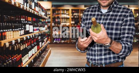 Mann in lässigen Kleidern, der Wein im Spirituosenladen auswählt. Der männliche Kunde hält zwei Weinflaschen in der Hand, während er in der Weinhandlung steht. Stockfoto