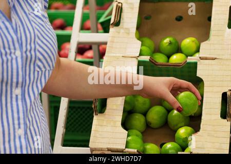 Die weibliche Hand nimmt gelbe, grüne Früchte in einem Supermarkt. Mädchen mit Kalk. Das Konzept der Fruchtwahl. Käuferkonzept. Kalkkästen Stockfoto