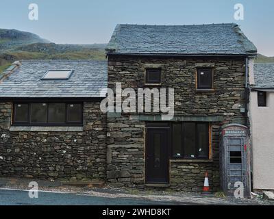 OD britischer Telefonkiosk grau lackiert, in einem Zustand der Verwüstung vor einem traditionellen Lakeland-Steingebäude. Die Telefonboxen sind normalerweise rot lackiert. Stockfoto