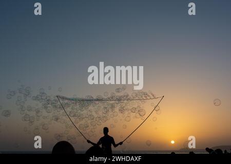 Ein Mann in der Silhouette macht viele Seifenblasen mit einem Seifenblasenstab, sie schweben am orangefarbenen Himmel bei Sonnenuntergang Stockfoto