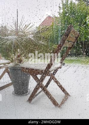 Blick durch die Regentropfen am Fenster auf die grünen Pflanzen und das Wetterphänomen Schnee im Mai in Berlin aufgrund des Klimawandels Stockfoto