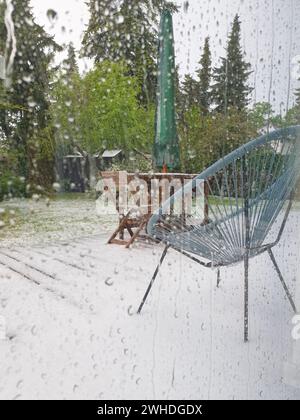 Blick durch die Regentropfen am Fenster auf die grünen Pflanzen draußen und das Wetterphänomen Schnee im Mai in Berlin aufgrund des Klimawandels Stockfoto