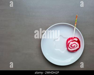 Ein rot-weißer Lutscher mit einem Stock liegt auf einem Teller vor einem hellgrauen Hintergrund Stockfoto