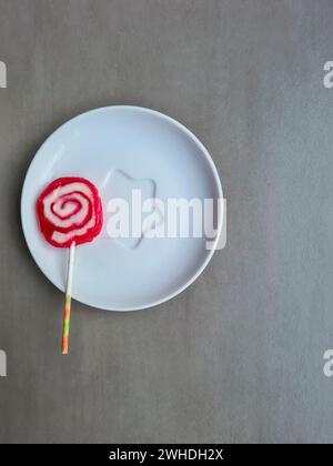 Ein rot-weißer Lutscher mit einem Stock liegt auf einem Teller vor einem hellgrauen Hintergrund Stockfoto